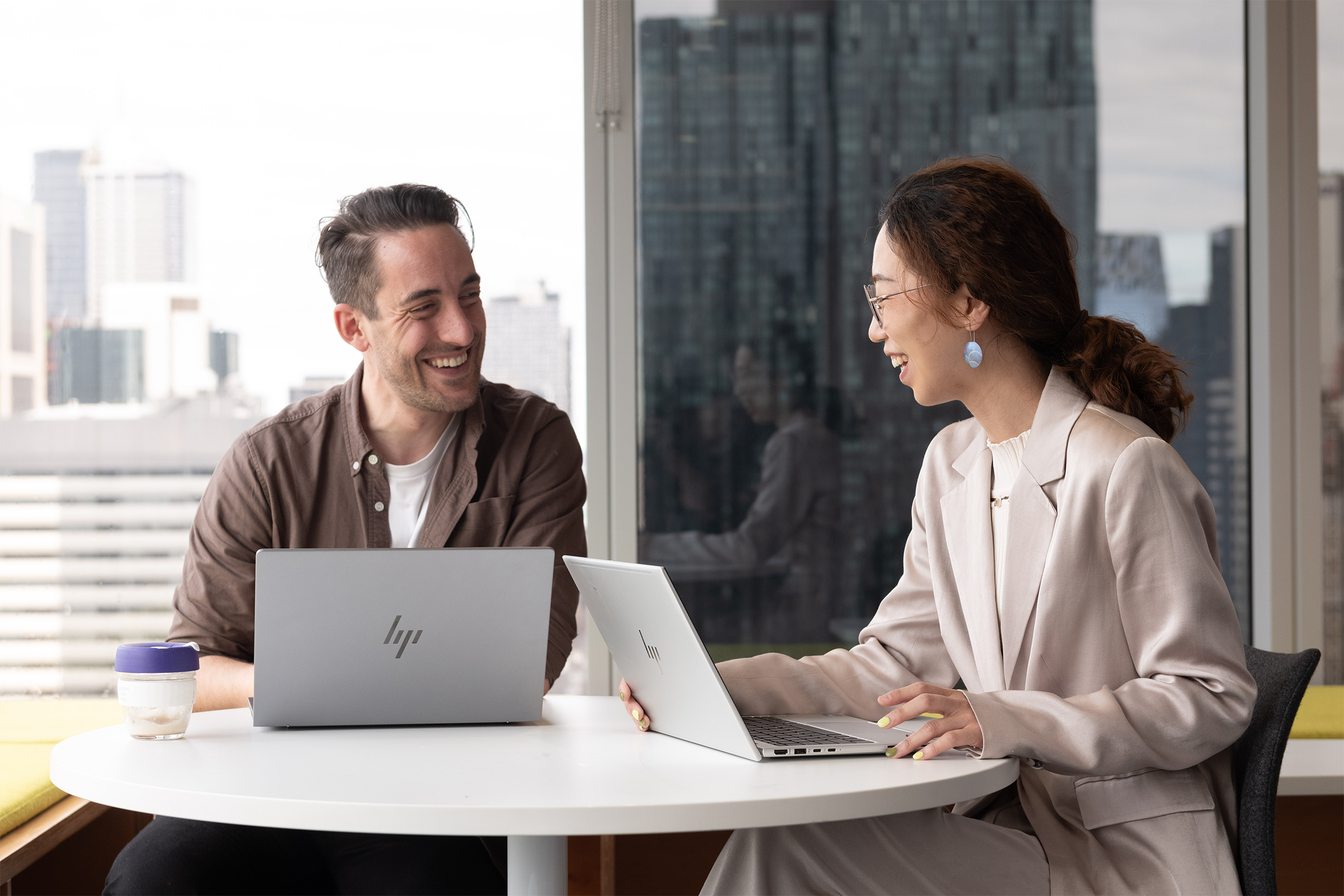 Two professionals sit together in an office, smiling and discussing content displayed on a laptop.
